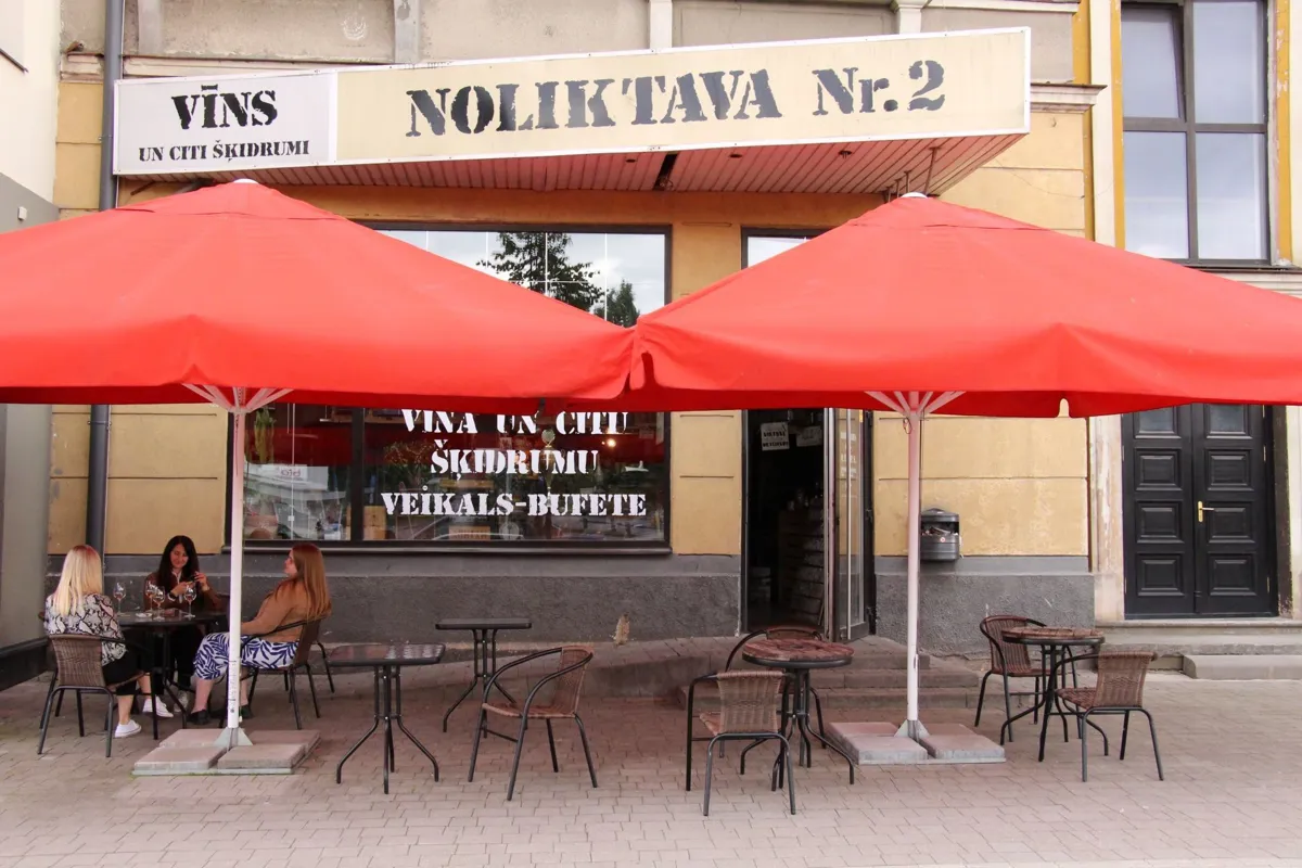 Noliktavas Nr. 2 fasāde Lielajā ielā ar izkārtni „Vīns un citi šķidrumi” un vasaras terasi zem sarkaniem saulessargiem