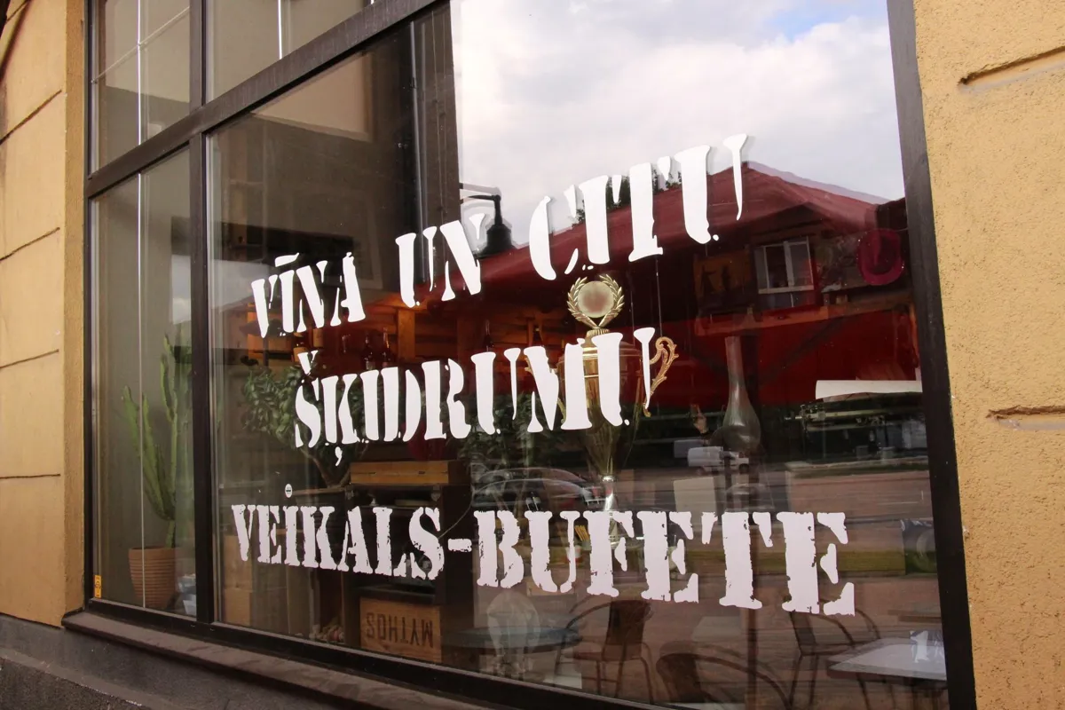 Skatloga uzraksts „Vīna un citu šķidrumu veikals-bufete” baltiem trafaretburtiem uz stikla